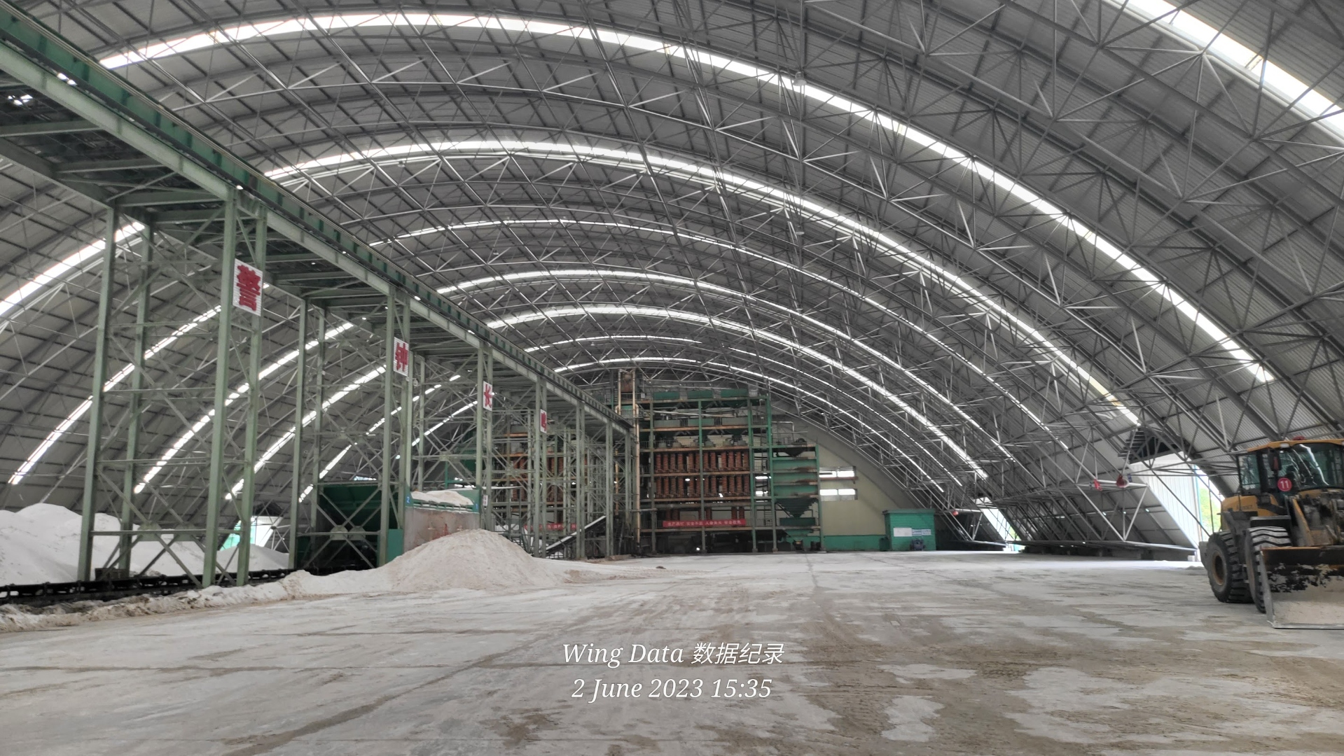 Interior of an arched steel-truss warehouse holding two white silica-sand stockpiles and a Volvo wheel loader.