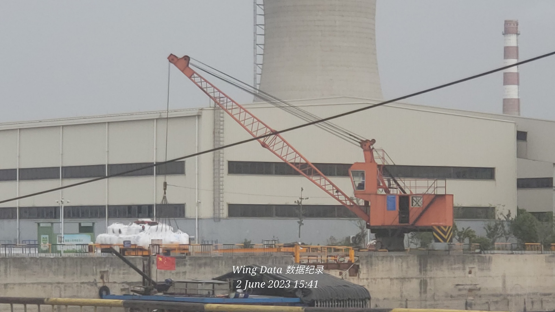 Riverside dock with an orange crawler crane loading white super-sacks of silica sand onto a river barge, cooling tower and striped stack in background.